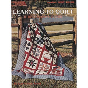 May include: A craft leaflet titled Learning to Quilt: A Beginner's Guide, featuring a quilt with red, white, and blue geometric patterns, including star and basket motifs, draped over a wooden fence in a grassy outdoor setting.
