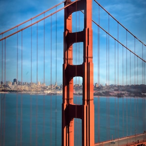 May include: A red steel suspension bridge with a large tower and a roadway. The bridge is over a body of water with a city skyline in the background.