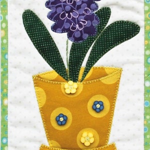 May include: A purple and green fabric hyacinth flower in a yellow pot with white polka dots and yellow, blue, and white buttons. The pot is sitting on a white background with a green and white checkered border.