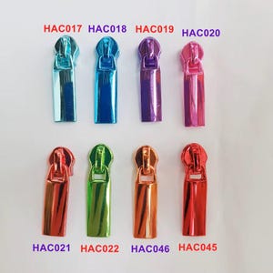 Könnte beinhalten: Zehn Reißverschluss-Zipper in verschiedenen Farben, darunter Blau, Lila, Pink, Rot, Orange und Grün. Die Reißverschluss-Zipper sind mit den Codes HAC017, HAC018, HAC019, HAC020, HAC021, HAC022, HAC046 und HAC045 gekennzeichnet.