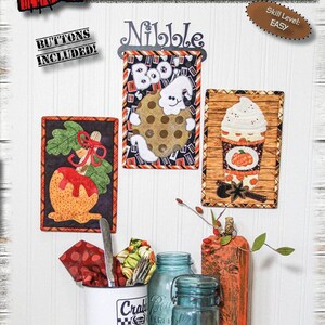 Könnte beinhalten: Eine herbstliche Präsentation mit Stoffkunstwerken und dekorativen Gegenständen. Die gerahmten Stoffkunstwerke sind ca. 15 cm x 23 cm groß. Die Präsentation umfasst Gläser, einen Becher und einen Weidenkorb. Der Text "Autumn Nibblers" ist sichtbar.