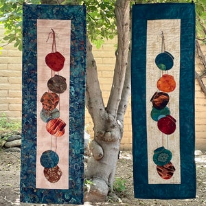 May include: Two vertical quilted wall hangings. Each features a central panel with a string of colorful, abstract shapes. The panels are framed by a teal fabric with a swirling pattern. The shapes are in shades of red, brown, and teal.