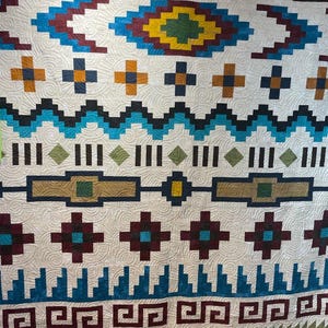 May include: A white quilt with a geometric pattern in shades of blue, green, red, and brown. The pattern is reminiscent of Native American art.