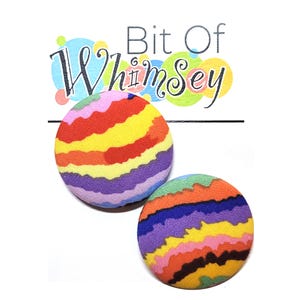 Puede incluir: Dos botones redondos coloridos con un patrón de rayas de arcoíris. Los botones están hechos de tela y tienen un fondo blanco.