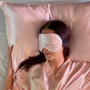 Puede incluir: Una persona con una m&aacute;scara de dormir de sat&eacute;n rosa y una bata de sat&eacute;n rosa est&aacute; acostada sobre una almohada de sat&eacute;n rosa.