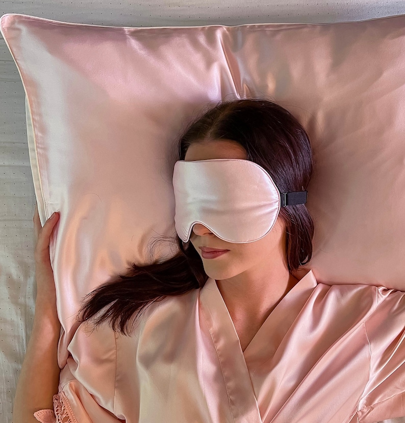 Puede incluir: Una persona con una m&aacute;scara de dormir de sat&eacute;n rosa y una bata de sat&eacute;n rosa est&aacute; acostada sobre una almohada de sat&eacute;n rosa.