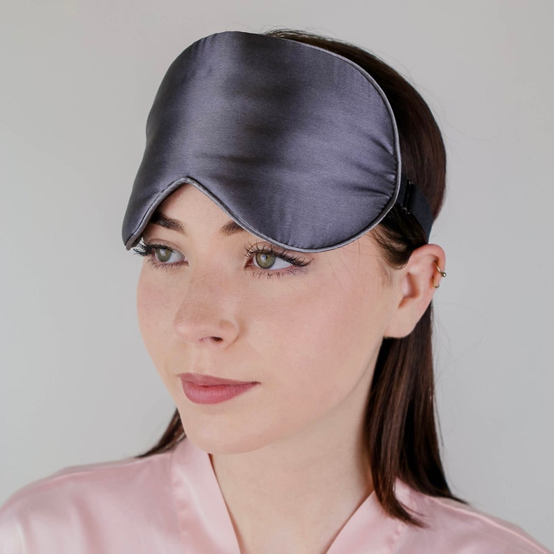 Puede incluir: Una m&aacute;scara de dormir de seda gris con una forma contorneada para bloquear la luz. La m&aacute;scara la lleva puesta una persona con cabello casta&ntilde;o.