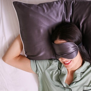 Puede incluir: Una persona con una camisa verde y una m&aacute;scara para dormir gris est&aacute; acostada sobre una almohada blanca con una funda de almohada de sat&eacute;n gris.