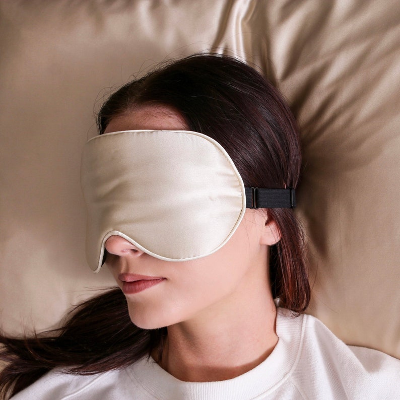 Puede incluir: Una persona lleva una m&aacute;scara de dormir de seda blanca con una banda el&aacute;stica negra. La m&aacute;scara cubre sus ojos y est&aacute; tumbada sobre una almohada beige.