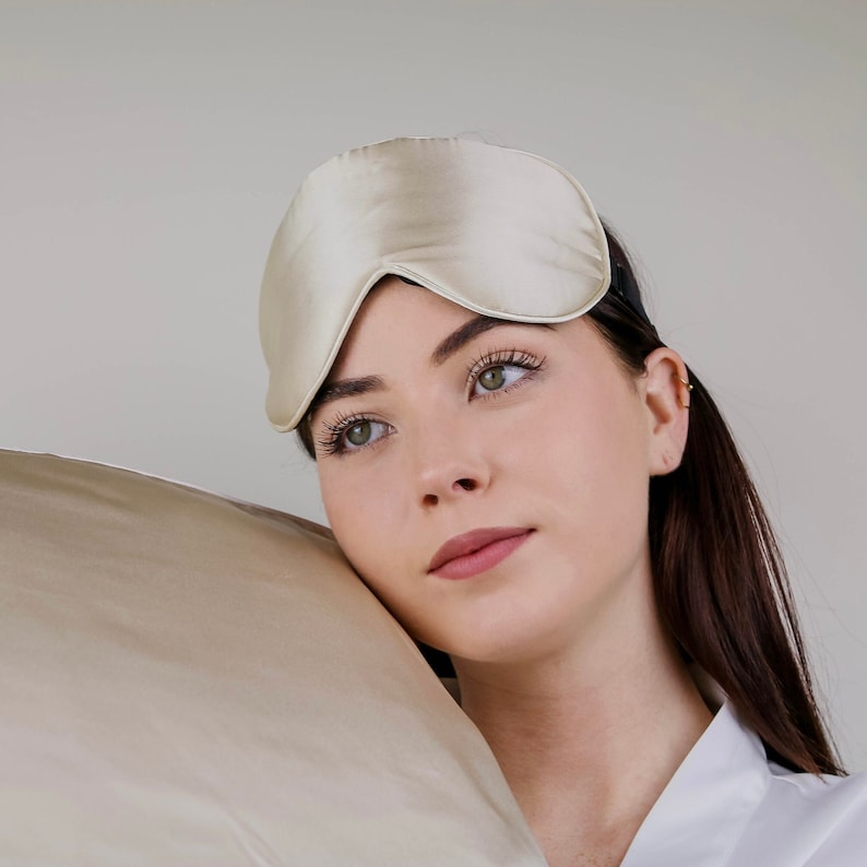 Puede incluir: Una persona con una m&aacute;scara de dormir de seda beige y una bata blanca, descansando la cabeza sobre una almohada de seda beige.