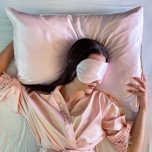 Puede incluir: Una persona con una bata de sat&eacute;n rosa y una m&aacute;scara para dormir de sat&eacute;n rosa est&aacute; acostada sobre una almohada de sat&eacute;n rosa.