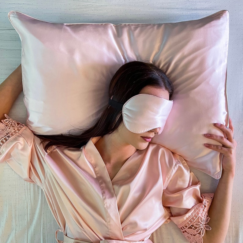 Puede incluir: Una persona con una bata de sat&eacute;n rosa y una m&aacute;scara para dormir de sat&eacute;n rosa est&aacute; acostada sobre una almohada de sat&eacute;n rosa.