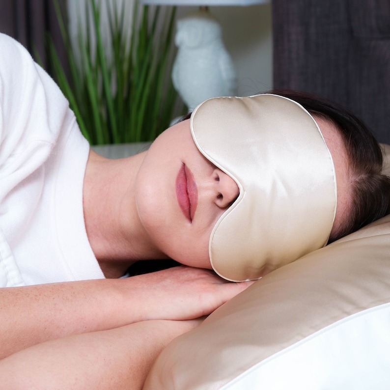 Puede incluir: Una persona durmiendo con una m&aacute;scara de dormir de seda beige sobre los ojos. La persona est&aacute; acostada sobre una almohada blanca y beige.