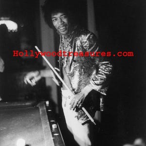 May include: A black and white photo of a man playing pool. He is wearing a sequined jacket and white pants. He is holding a pool cue and looking at the camera. The photo is taken from a low angle and the pool table is in the foreground.