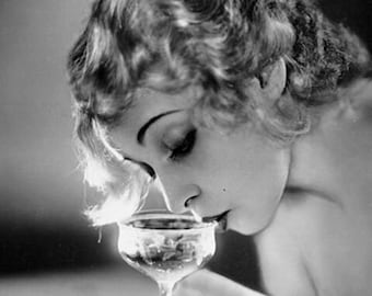 Op de afbeelding: Zwart-witfoto van een vrouw met golvend haar, die naar beneden kijkt naar een glazen steelglas gevuld met vloeistof. De tekst "Hollywoodtreasures.com 16" x 20" " is zichtbaar onderaan de afbeelding.