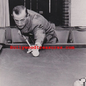 May include: Black and white photograph of a man playing billiards. The man is wearing a suit and is focused on the shot. A pool cue is in his hand, and a white ball is in front of him. The pool table is green.