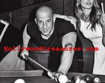 May include: A black and white photograph of a man playing pool. He is wearing a black t-shirt and jeans. He is leaning over the pool table, aiming his cue stick at a white billiard ball. A woman in a white shirt is standing behind him, watching him play. The text "Hollywoodtreasures.com" is visible in the image.