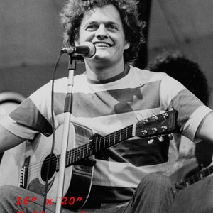 May include: A black and white photograph of a man with curly hair playing a guitar. He is wearing a striped shirt and holding a microphone. The guitar is a six-string acoustic guitar. The photo is 16 inches by 20 inches.