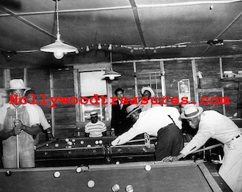 May include: A black and white photograph showing a group of men playing pool in a dimly lit room. The men are wearing hats and casual clothing. There are two pool tables in the room, and the men are gathered around one of them. The photo is taken from a low angle, and the men's faces are obscured by the pool table.