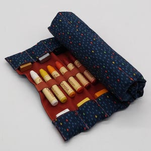 May include: A navy blue crayon roll with a colourful raindrop pattern, open to reveal crayons in various colours. The interior is red, with brown straps holding the crayons in place. The crayons are labelled with the word "CAMAR" and "MALSTIFT".