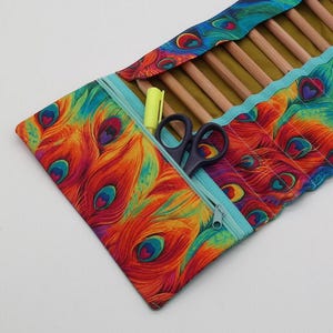 Könnte beinhalten: Eine türkisfarbene Reißverschlusstasche mit einem Pfauenfeder-Druckmuster. Die Tasche ist geöffnet und zeigt eine Reihe von Holzstiften, eine Schere und einen Textmarker.