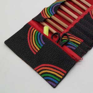 May include: A black fabric knitting needle case with a red interior and a rainbow pattern. The case is open, revealing wooden knitting needles, a yellow highlighter, and black scissors. The case has a red zipper.