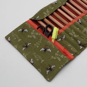 May include: A green fabric pencil case with a bee and floral pattern. The case has an orange zipper and interior, and contains pencils, scissors, and a yellow highlighter. The case is open, revealing the contents.