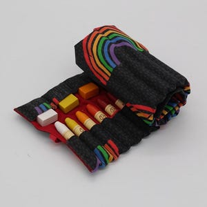May include: A rainbow-patterned fabric roll-up case with a black background and red trim. The case is open to reveal a set of crayons and a white eraser.