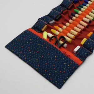 May include: A navy blue, roll-up art supply case with a red interior and orange zipper. The case is filled with various art supplies, including crayons, pastels, and a pair of black scissors. The exterior has a pattern of small, colorful dots.