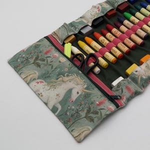 May include: A green fabric pencil case with a unicorn and floral pattern, open to reveal crayons, scissors, and erasers. The case has a red zipper and a dark green interior. The crayons are various colors.