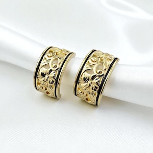 May include: A pair of gold-colored hoop earrings with a floral filigree design. The earrings have a black border and are displayed on a white satin surface. The earrings are a jewelry item.