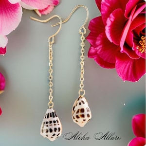 May include: Gold chain earrings with a seashell pendant. The seashells are white with brown markings. The earrings are hanging against a background of pink flowers.