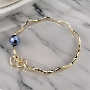 May include: A gold-toned bangle bracelet with a textured, wavy design. The bracelet features a single dark gray pearl and two interlocking heart charms.