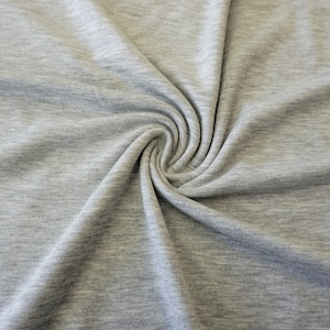 May include: A close-up shot of gray fabric with a subtle heathered texture. The material is gathered and swirled, creating soft folds and shadows. The fabric appears to be a knit or jersey type, suitable for clothing.
