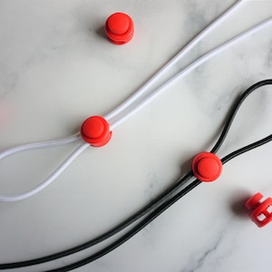 May include: Close-up of red cord locks and elastic cords. One cord is white, the other is black. The red cord locks are round and cylindrical. The cords are looped and ready for use.