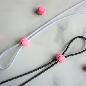 May include: Close-up of white and black elastic cords with pink cord locks and round stoppers. The cords are looped and arranged on a light-colored surface. The pink components add a pop of color.