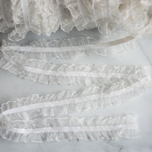 May include: White ruffled elastic trim. The trim is made of a sheer, delicate fabric with a ruffled edge. The elastic is visible on the back of the trim. The trim is rolled up in a spool and also laid out flat.