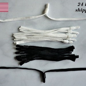 May include: An assortment of white and black elastic cords, likely for face masks or similar items. The image includes several cords with adjustable knots and metal fasteners. The American flag is in the upper left corner.
