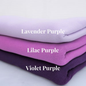 May include: A stack of folded knitwear in shades of purple: lavender, lilac, and violet. The image displays the color names on each layer. The fabric has a ribbed texture.