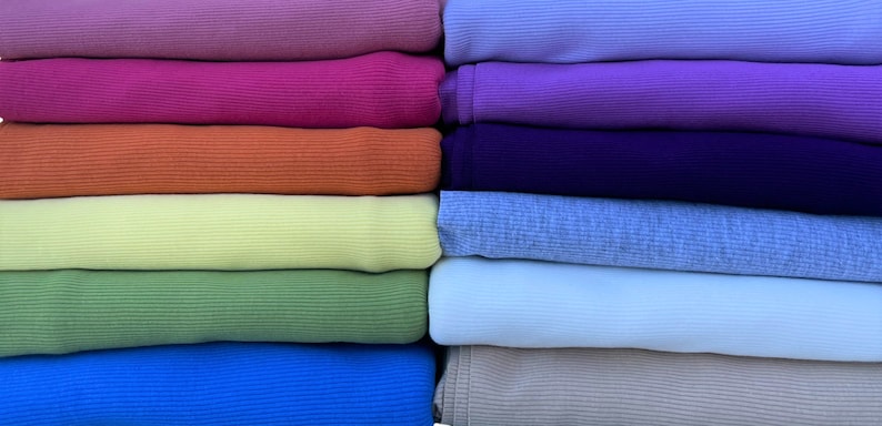 May include: A collection of folded fabric in various colors, including pink, orange, green, and blue. The fabrics appear to be a ribbed knit material, stacked neatly in rows. The colors range from light to dark shades.