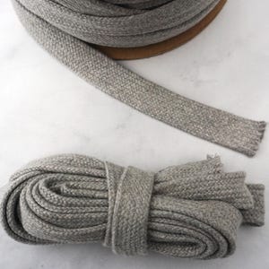 Könnte beinhalten: Eine Rolle grauer geflochtener Kordel mit einem abgewickelten Stück. Ein Bündel derselben Kordel ist ebenfalls sichtbar. Die Kordel hat ein strukturiertes Aussehen und wird wahrscheinlich für Bastel- oder Nähprojekte verwendet.