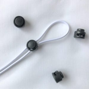 May include: Close-up of black cord locks and a white elastic cord against a white background. The cord is looped, and the locks are scattered around. The cord locks are small, round, and have a button-like appearance.