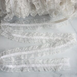 May include: White ruffled lace trim, ideal for sewing and crafting projects. The delicate lace is gathered along a thin, white band. A roll of the lace is visible in the background, with several lengths of the trim unrolled.