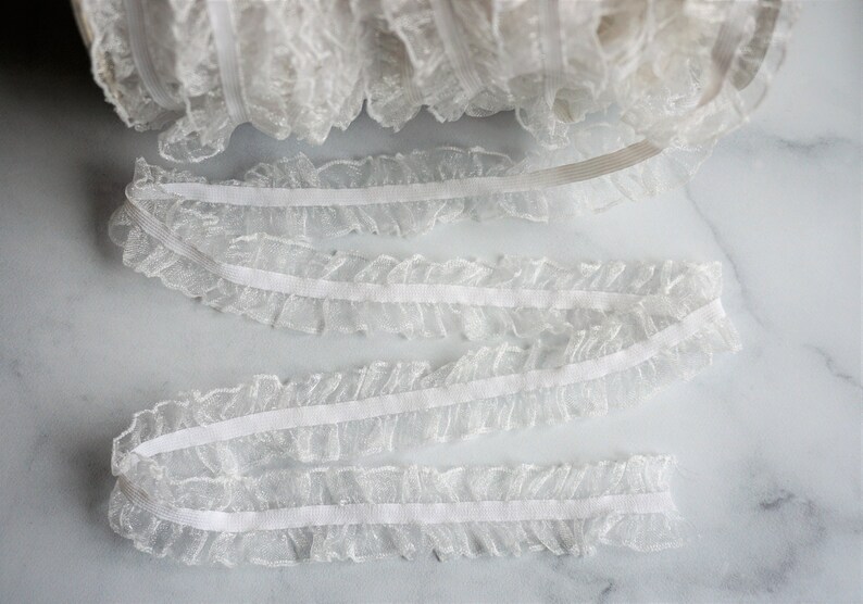 May include: White ruffled lace trim, ideal for sewing and crafting projects. The delicate lace is gathered along a thin, white band. A roll of the lace is visible in the background, with several lengths of the trim unrolled.