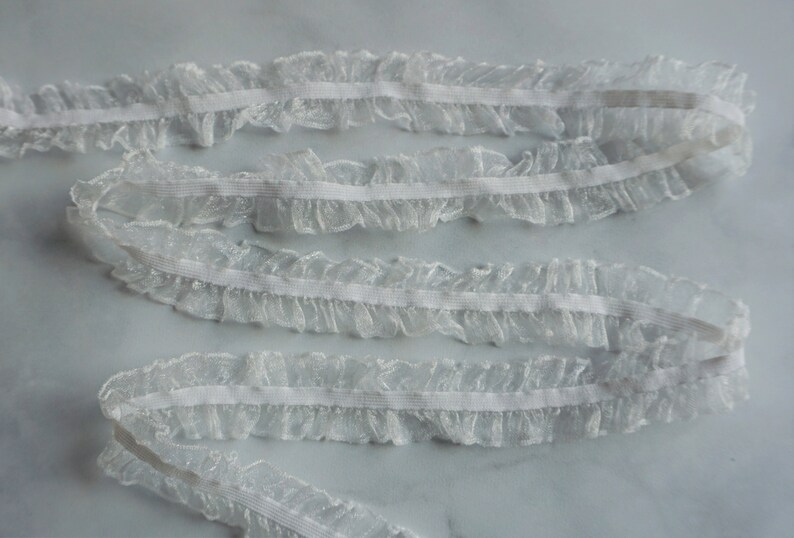 May include: White ruffled lace trim with an elastic band. The delicate, sheer fabric is gathered to create a ruffled edge. This trim is suitable for sewing, crafting, and adding a decorative touch to garments or accessories.