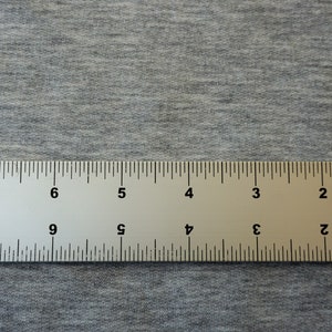 May include: A metal ruler with black markings and numbers, measuring in inches, lies on a gray fabric surface. The ruler is positioned horizontally across the image, providing a clear view of its markings.
