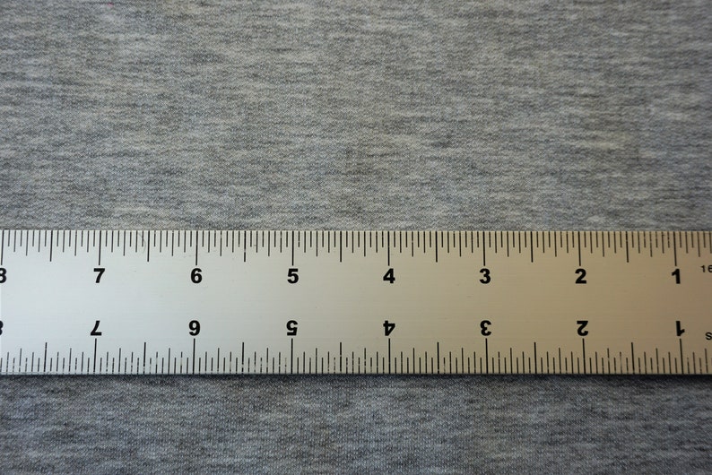 May include: A metal ruler with black markings and numbers, measuring in inches, lies on a gray fabric surface. The ruler is positioned horizontally across the image, providing a clear view of its markings.