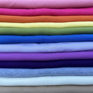 May include: A stack of colorful ribbed knit fabric in various shades, including pink, orange, yellow, green, blue, purple, and beige. The fabric is neatly folded, showcasing the texture and range of colors available for clothing or craft projects.