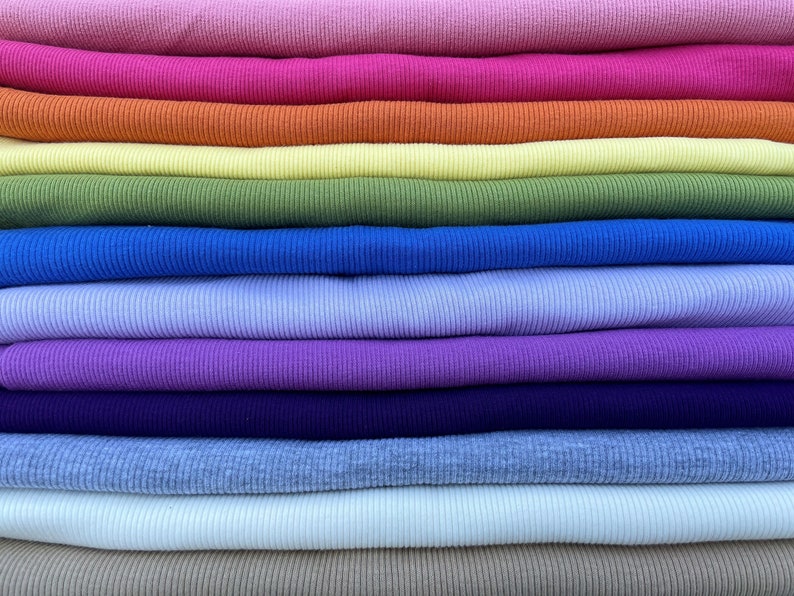 May include: A stack of colorful ribbed knit fabric in various shades, including pink, orange, yellow, green, blue, purple, and beige. The fabric is neatly folded, showcasing the texture and range of colors available for clothing or craft projects.