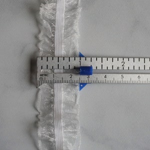 May include: Close-up of white lace trim with a ruffled edge, measured with a ruler. The trim is approximately 1 inch wide. The ruler shows both inches and centimeters for accurate measurement.